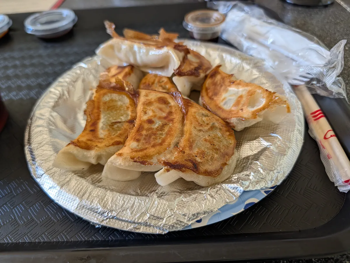 Eight pan fried dumplings on a foil plate with dipping sauce cups and wrapped chopsticks