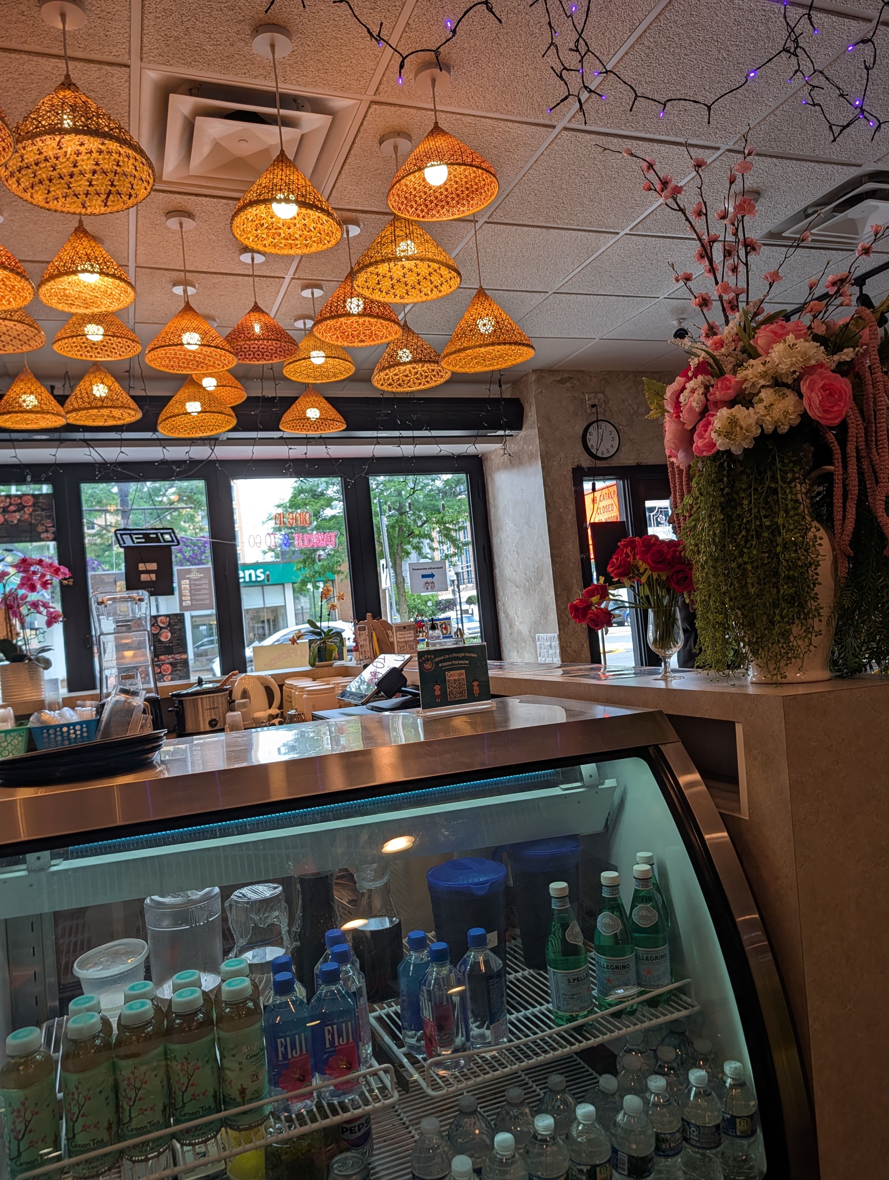 Kacha Thai Market front counter under a cluster of warm woven rattan lantern shades, with a drink cooler and fresh flowers