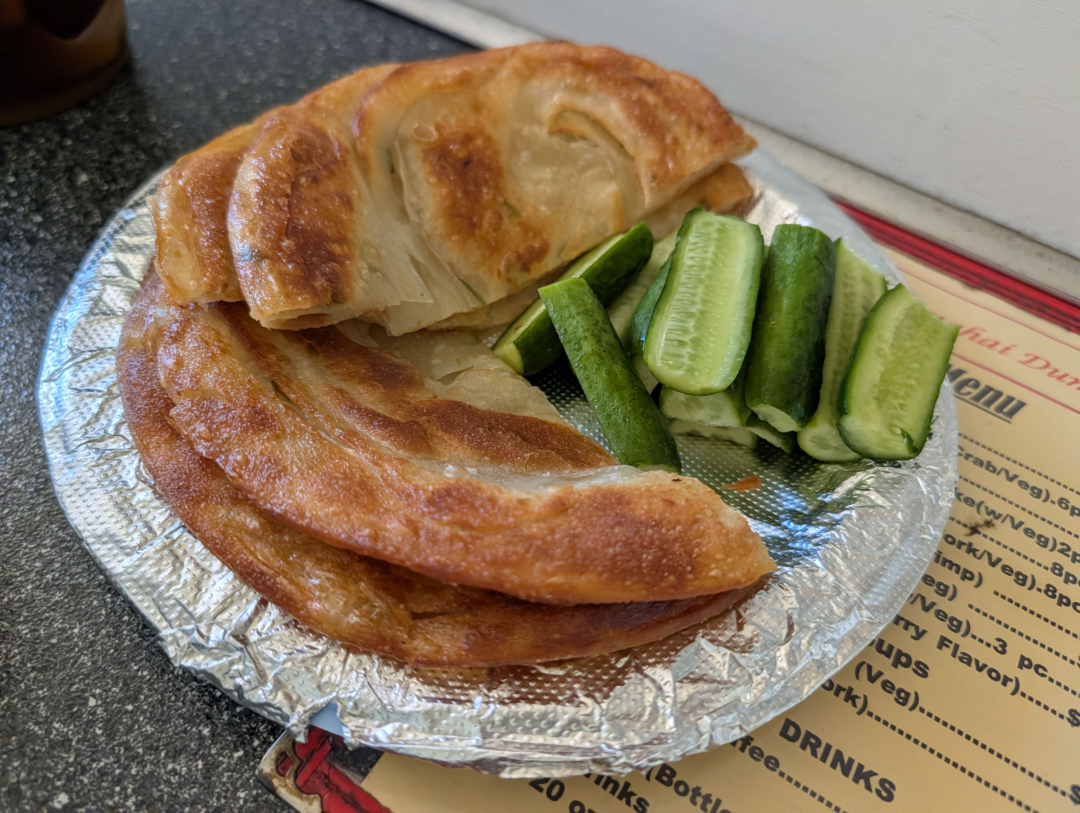 Two pieces of flaky green onion pancake on a foil plate with fresh cucumber spears