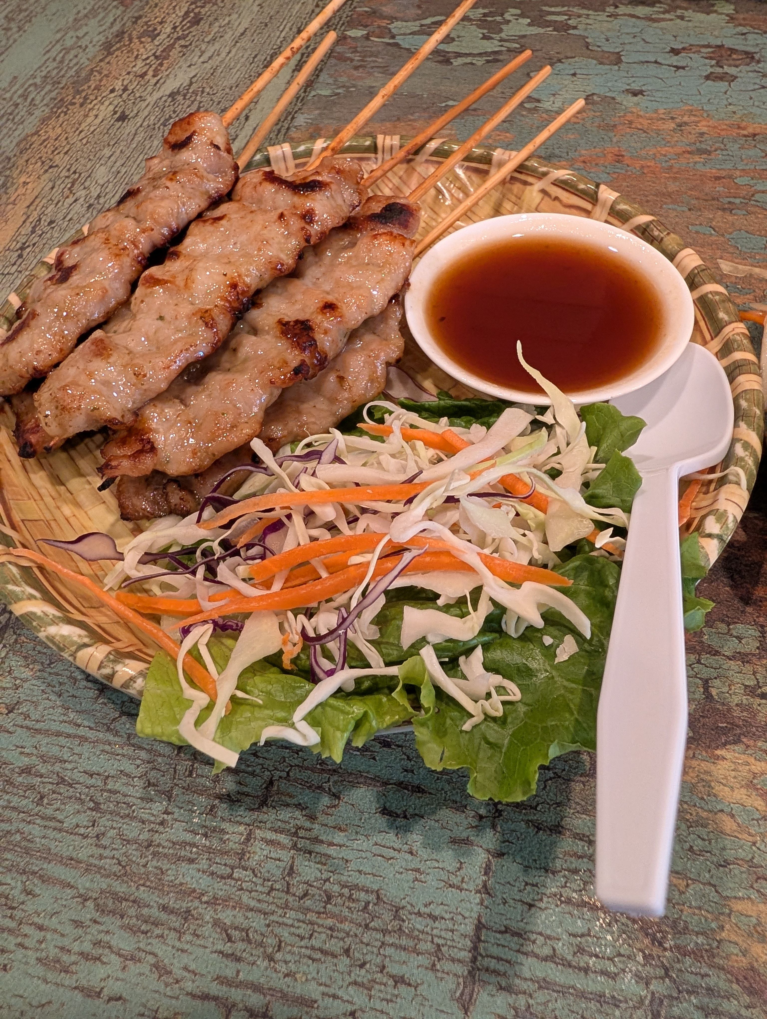 Grilled pork skewers on a woven bamboo plate with shredded cabbage and carrot slaw and a cup of dipping sauce