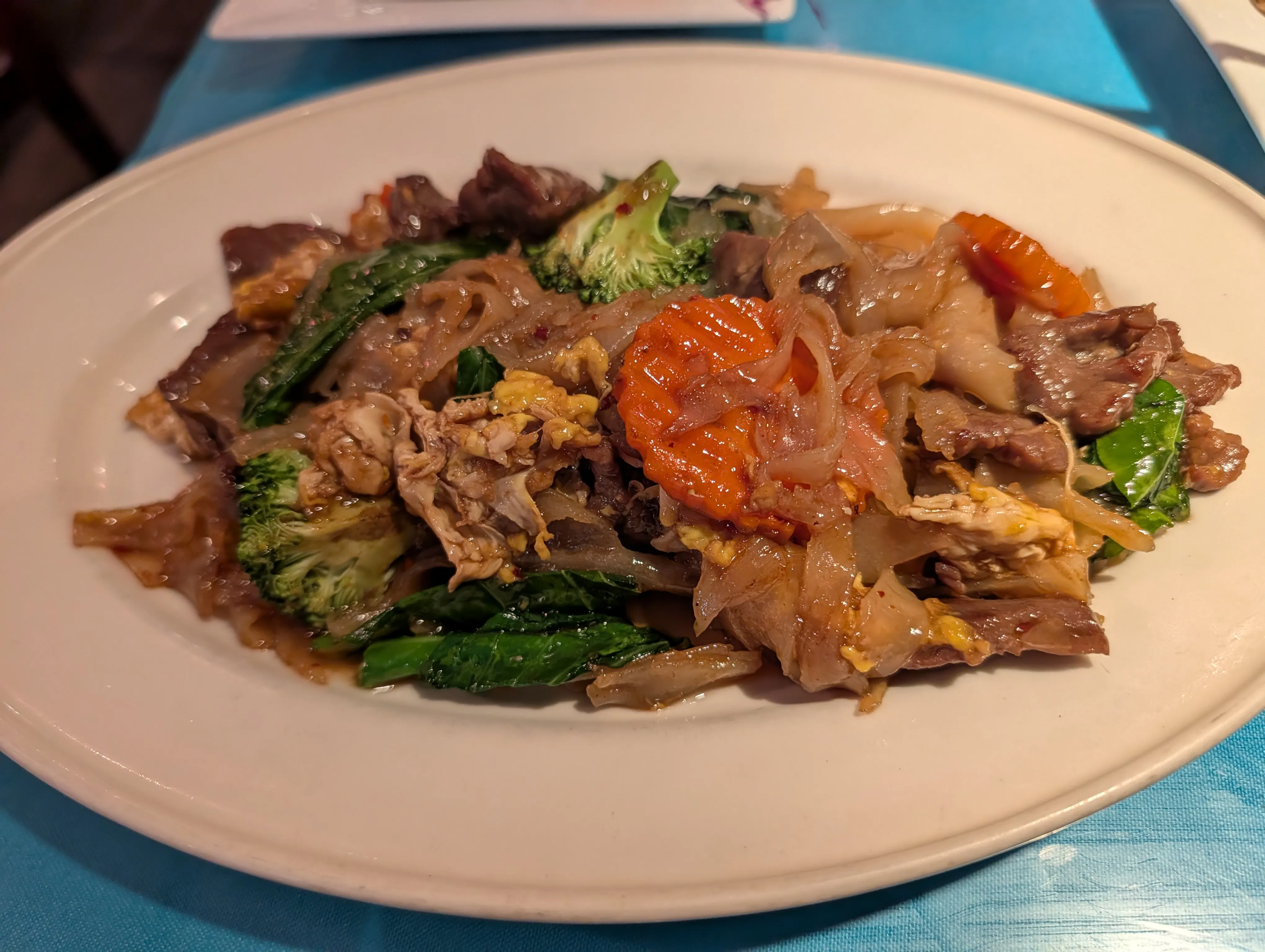 Wide rice noodles with beef, broccoli, carrot and Thai basil on a white oval plate