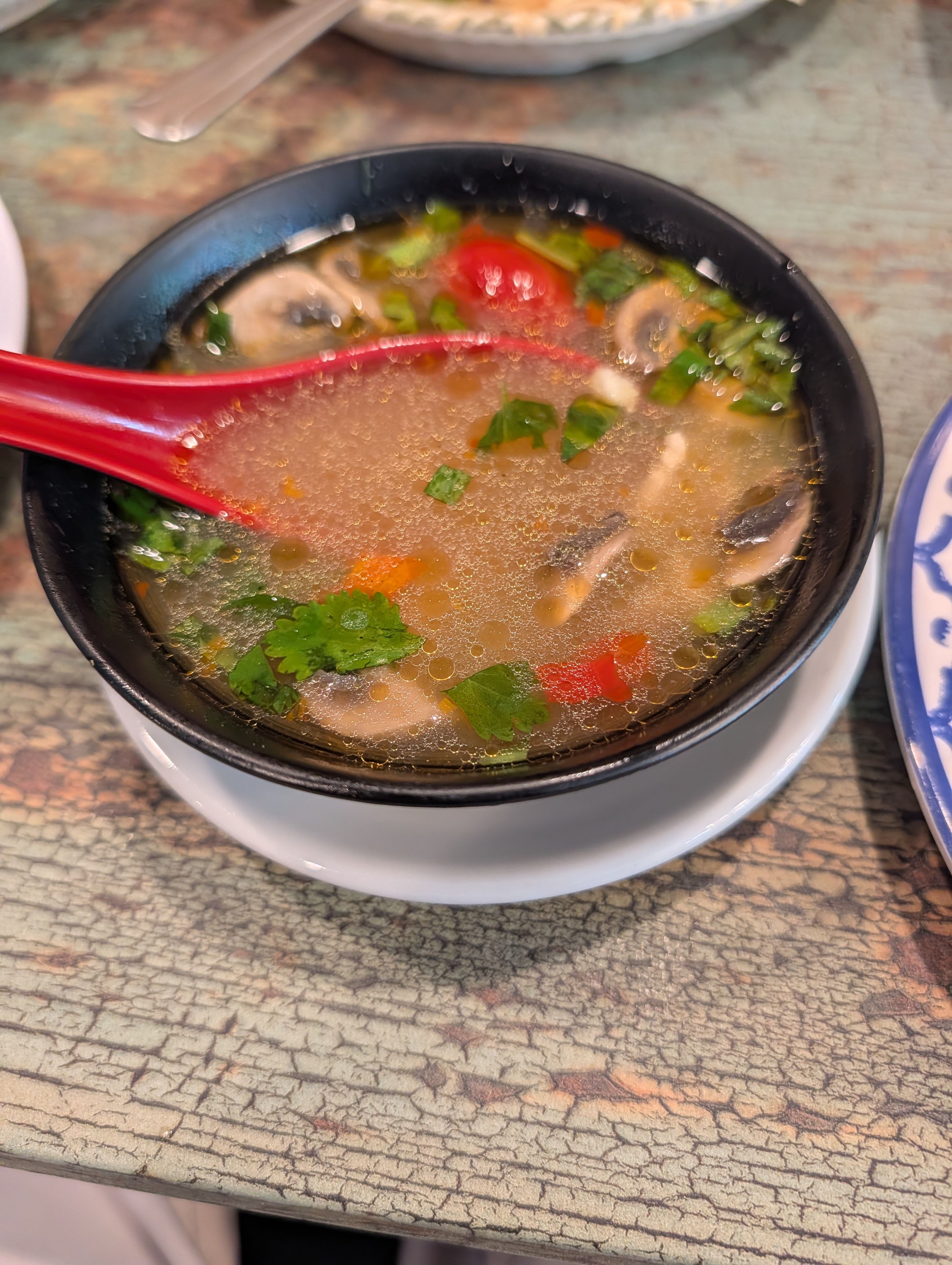 Clear tom yum soup with mushrooms, tomato, cilantro, and chili in a black bowl with a red soup spoon