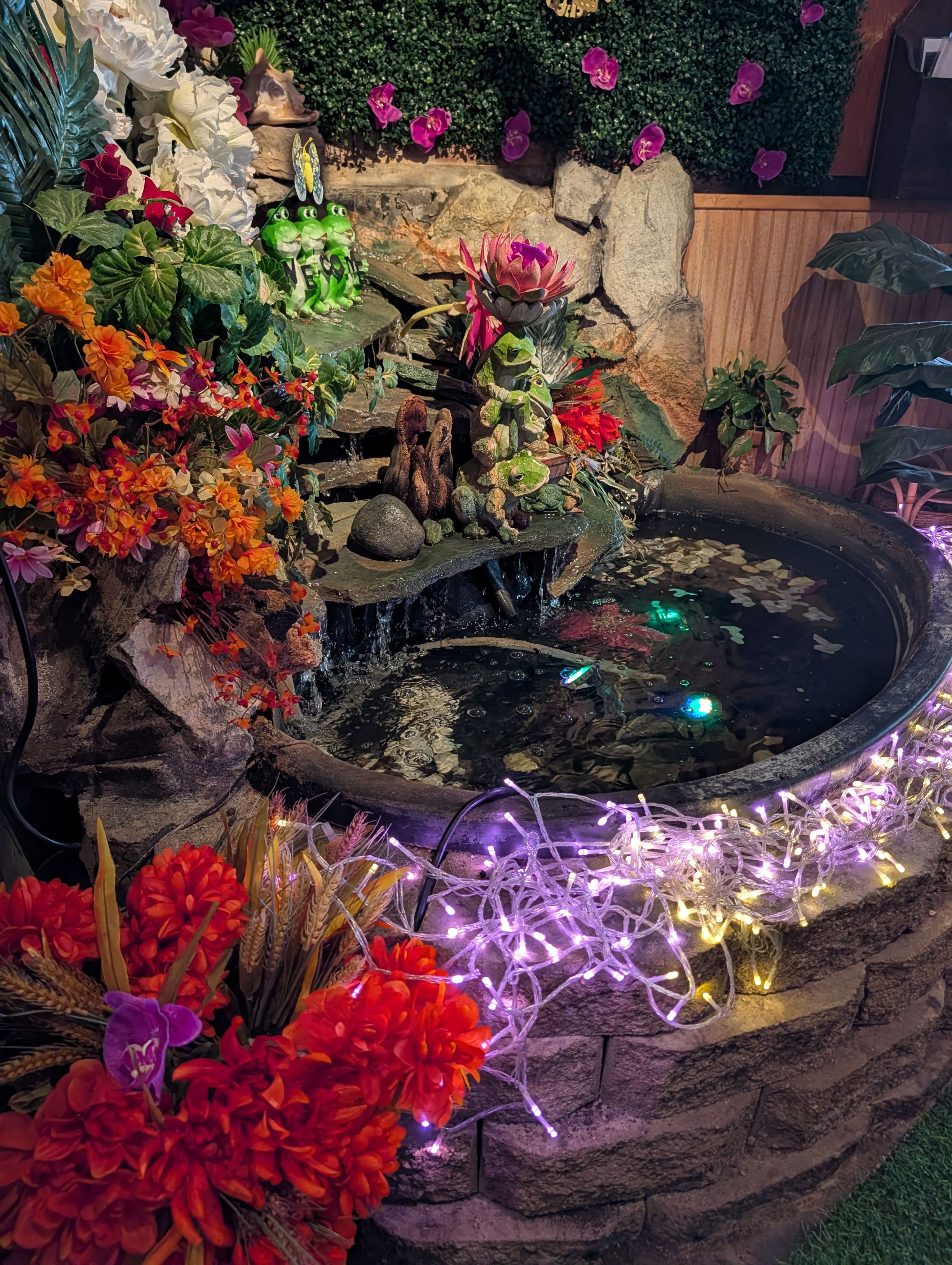 An indoor rock waterfall and pool decorated with artificial orchids, ceramic frogs and string lights